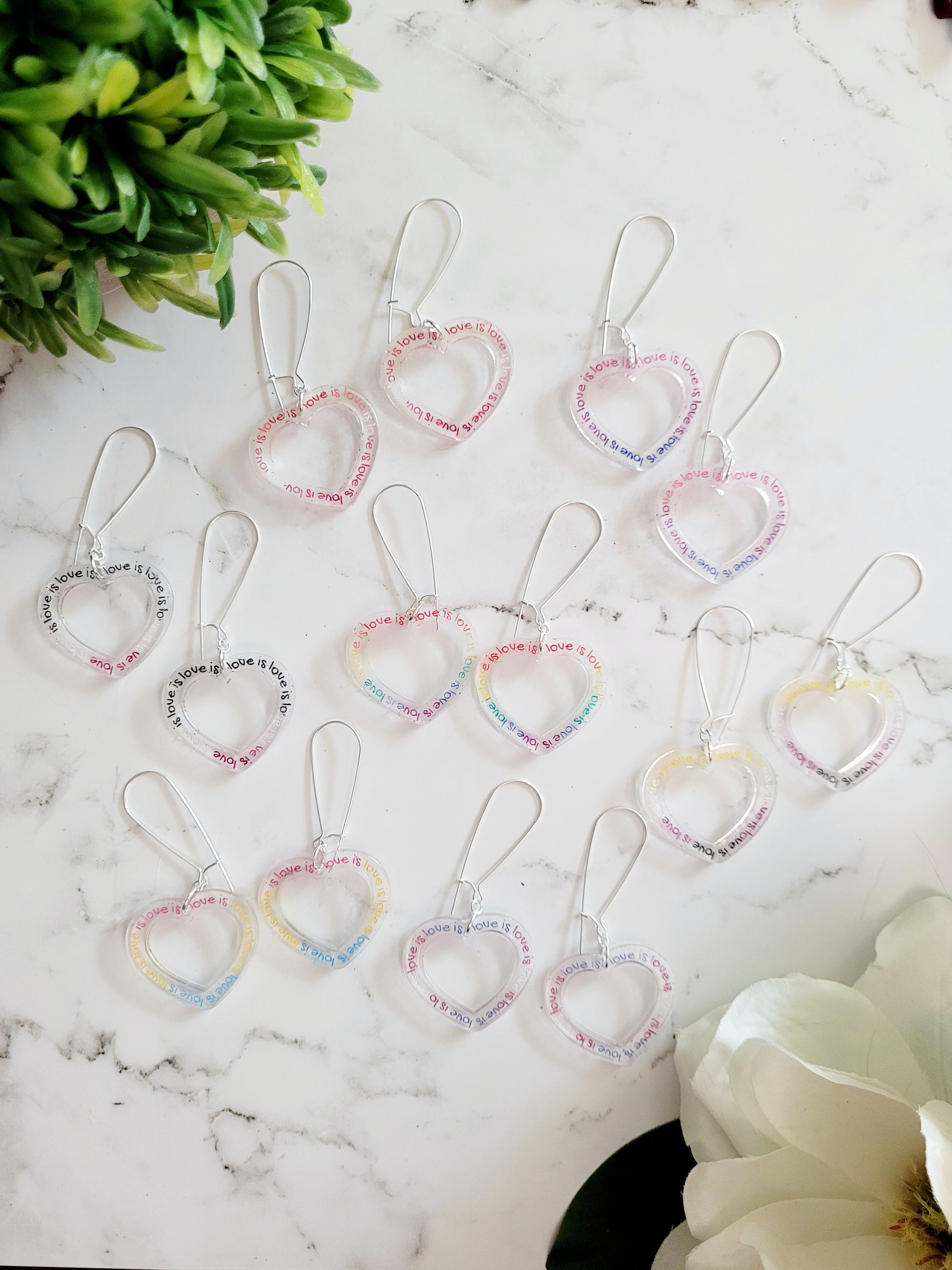 7 different Pride Flags earrings in the shape of a clear heart with Love is Love etched. 