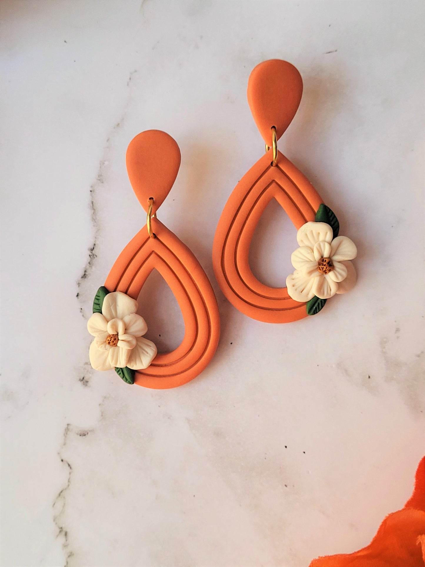 close up of orange teardrop earrings with a white magnolia flower sitting on a marble background.