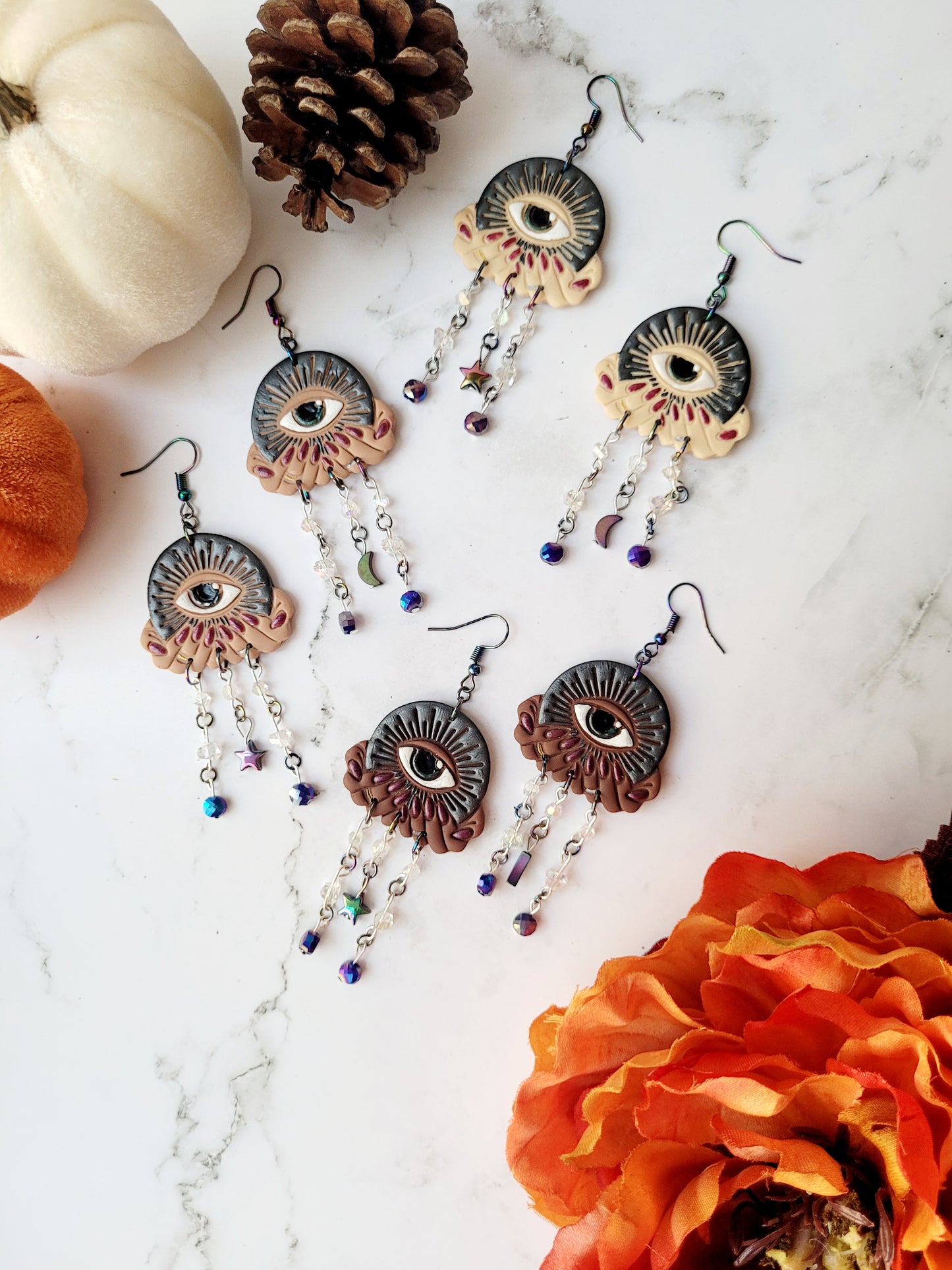 All seeing eye earrings in three skin tones on a white marble background with pumpkins and flowers. 