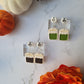 green and black tea bag shaped dangle earrings on a marble background with fall foliage. 