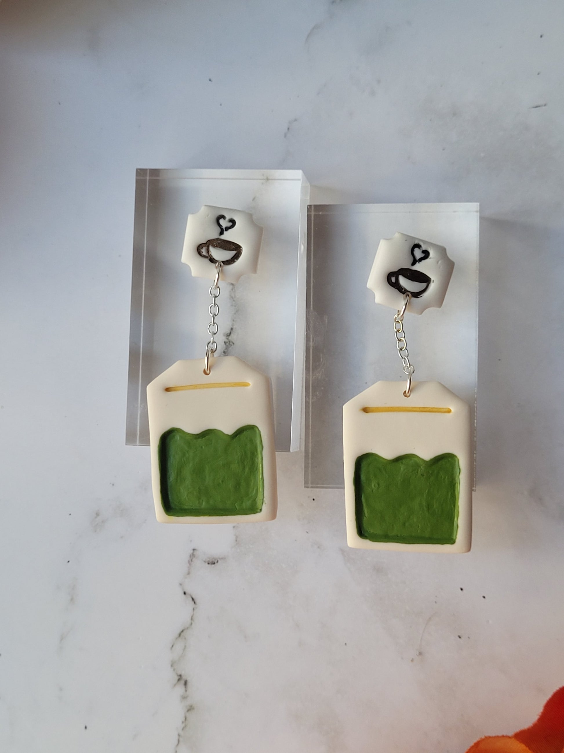 close up of green tea bag shaped dangle earrings on a marble background with fall foliage.