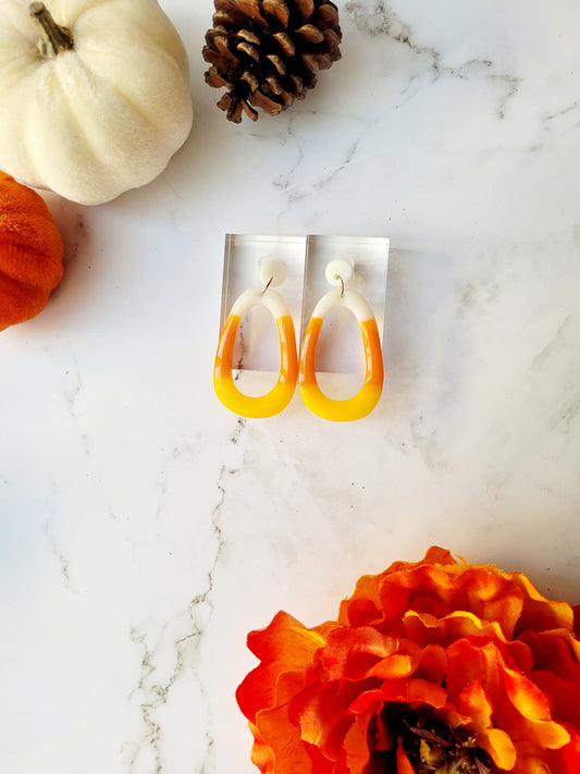 candy corn colored dangle earrings on a white marble background surrounded by fall foliage. 