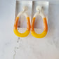 close up of candy corn colored dangle earrings on a white marble background surrounded by fall foliage.