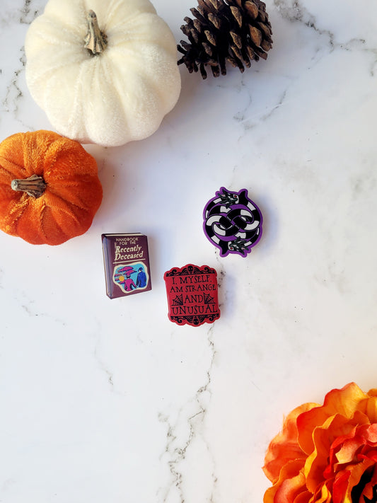 3 Beetlejuice themed brooches on a white marble background surrounded by fall foliage. 