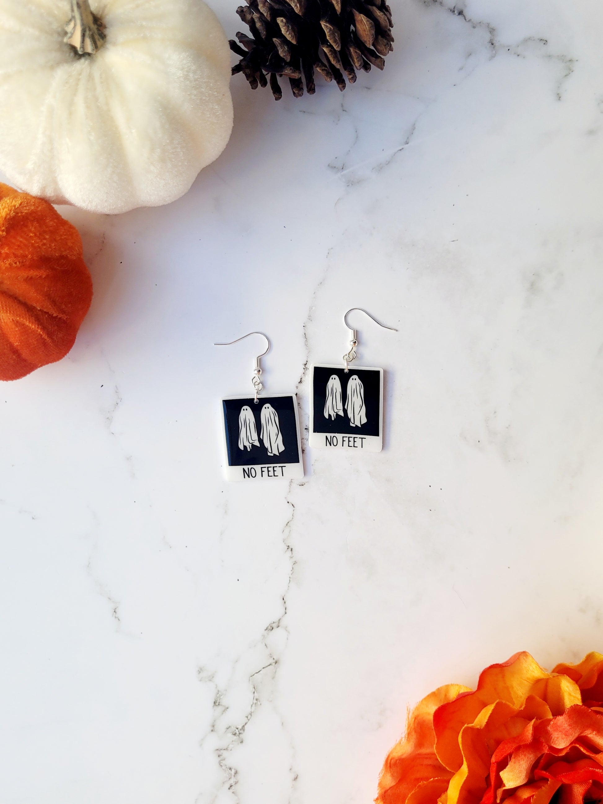 Beetlejuice inspired "no feet" ghost polaroid earrings on a marble background with fall foliage. 