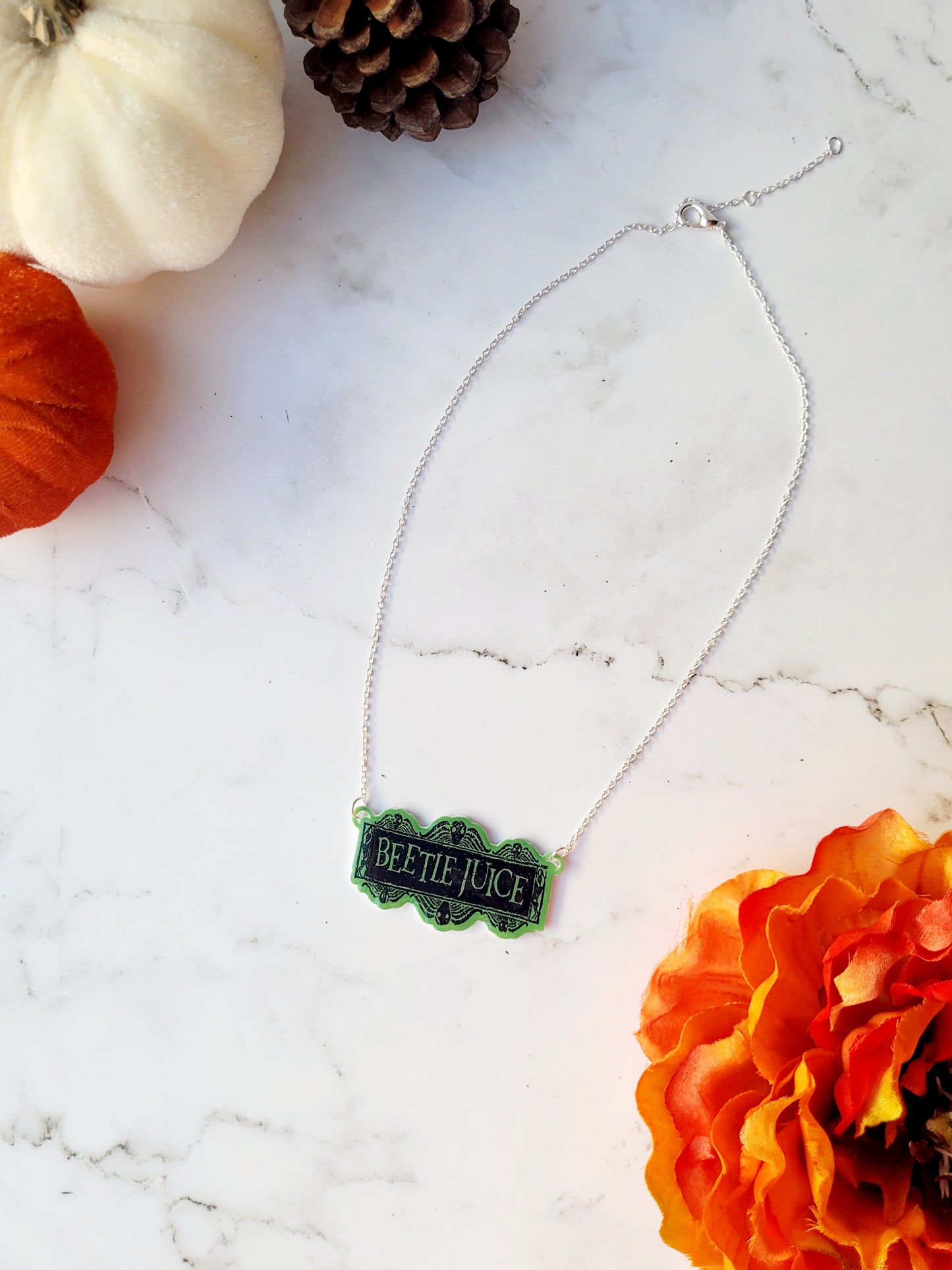 Beetlejuice logo necklace on a white marble background surrounded by fall foliage. 