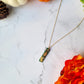  Green bar shaped pendant with metallic acorns and oak leaves. on a gold chain. Sitting on a white marble background with pumpkins and flowers in the background. 