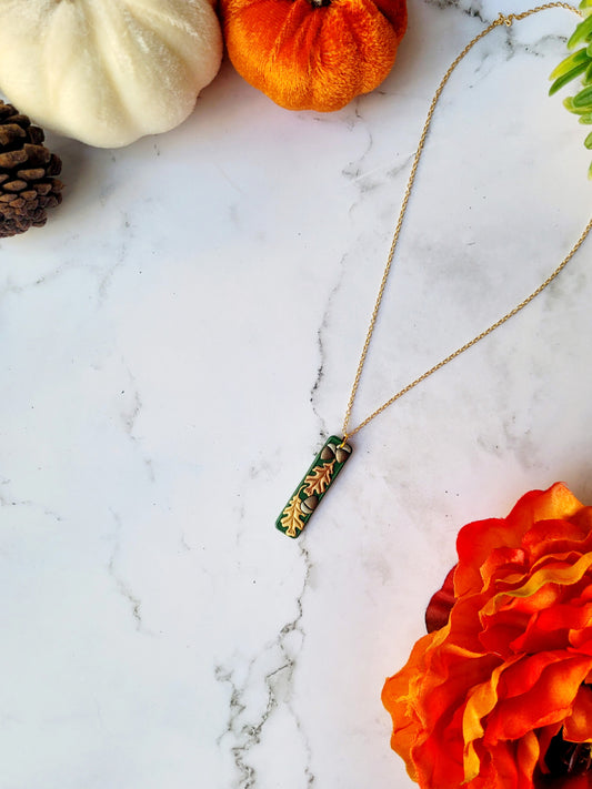  Green bar shaped pendant with metallic acorns and oak leaves. on a gold chain. Sitting on a white marble background with pumpkins and flowers in the background. 