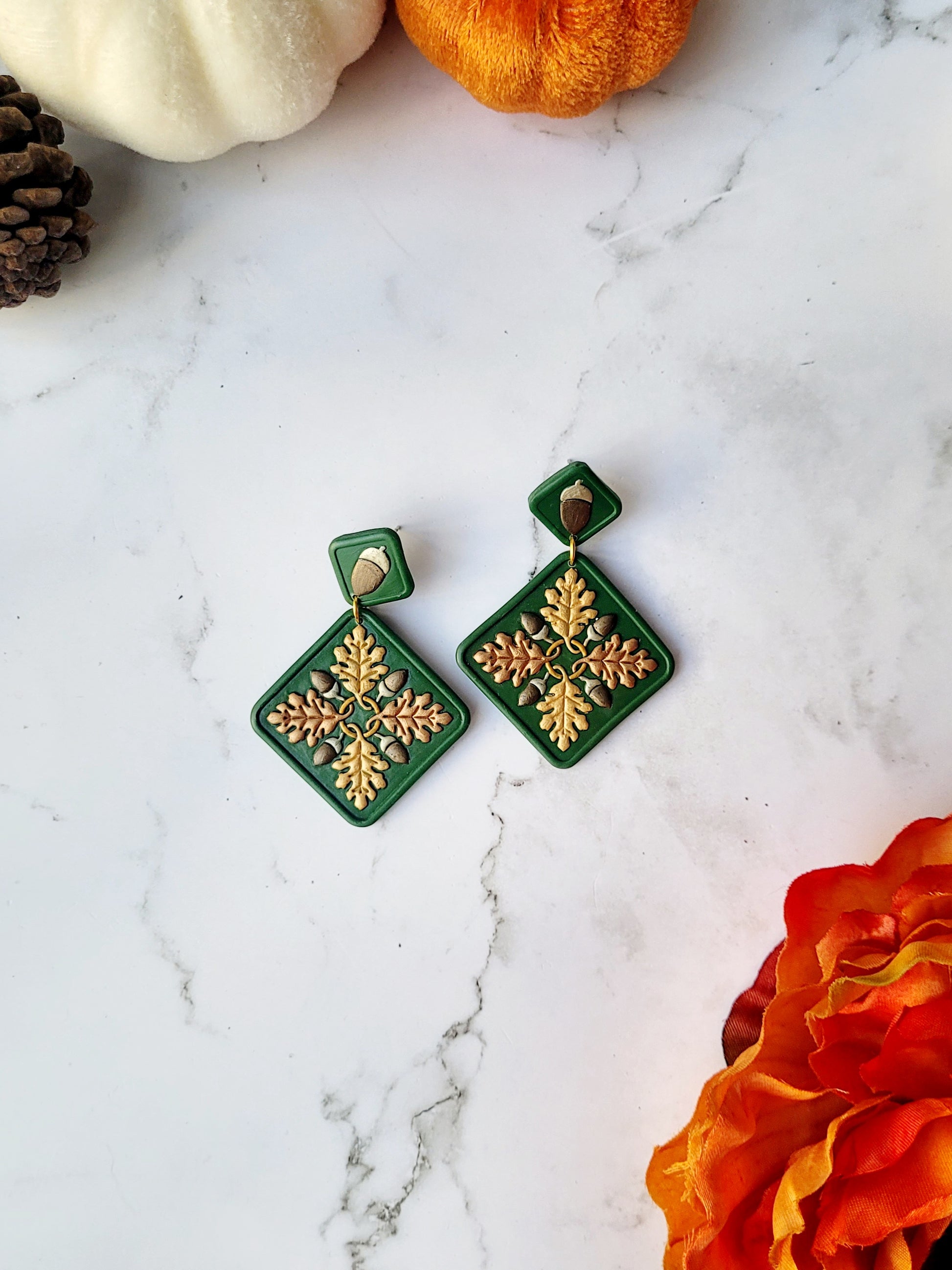 Green diamond shaped earrings with metallic acorns and oak leaves. Sitting on a white marble background with pumpkins and flowers in the background. 