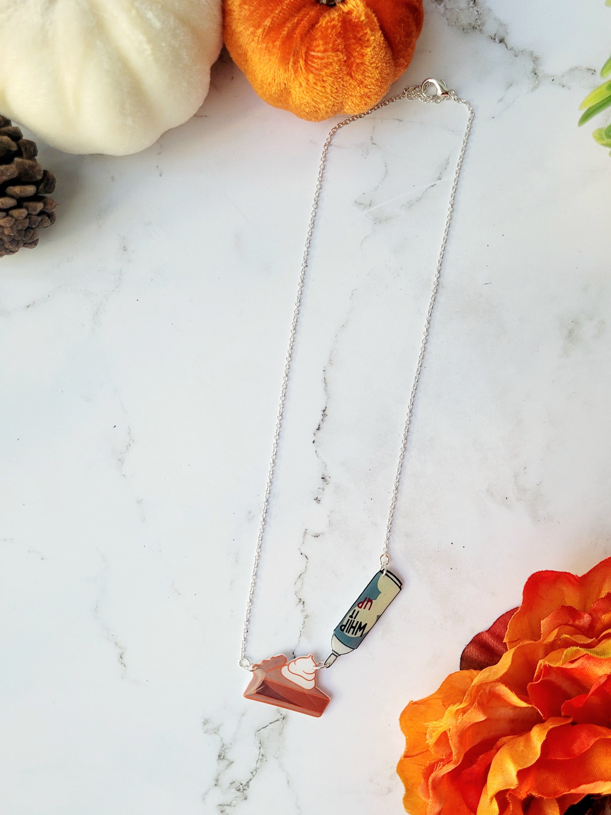 Pumpkin pie slice necklace with a can of whipped cream on a marble background surrounded by fall foliage. 