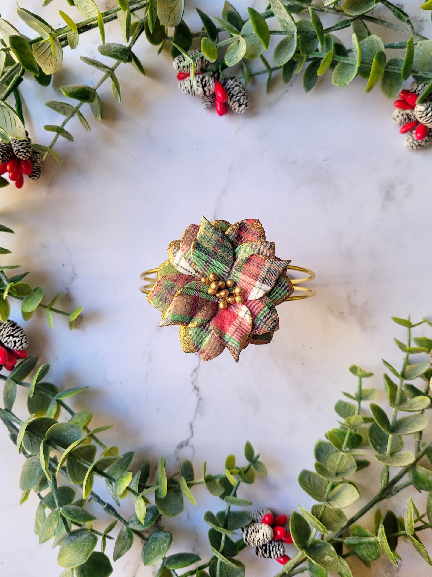 Christmas themed plaid poinsettia bracelet on a marble background surrounded by foliage. 