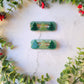 Green glitter hair clips with christmas tree charms on a white marble background with foliage. 