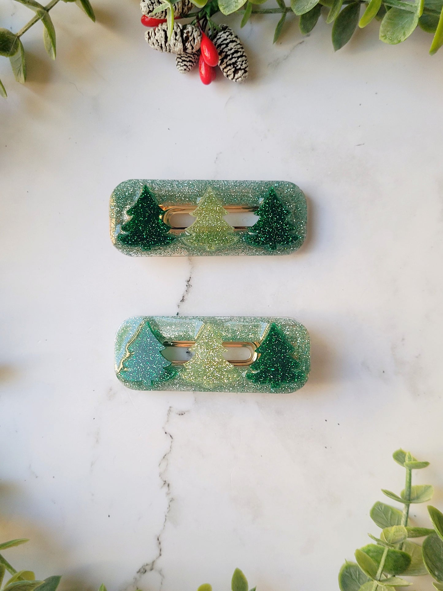 close up of Green glitter hair clips with christmas tree charms on a white marble background with foliage.
