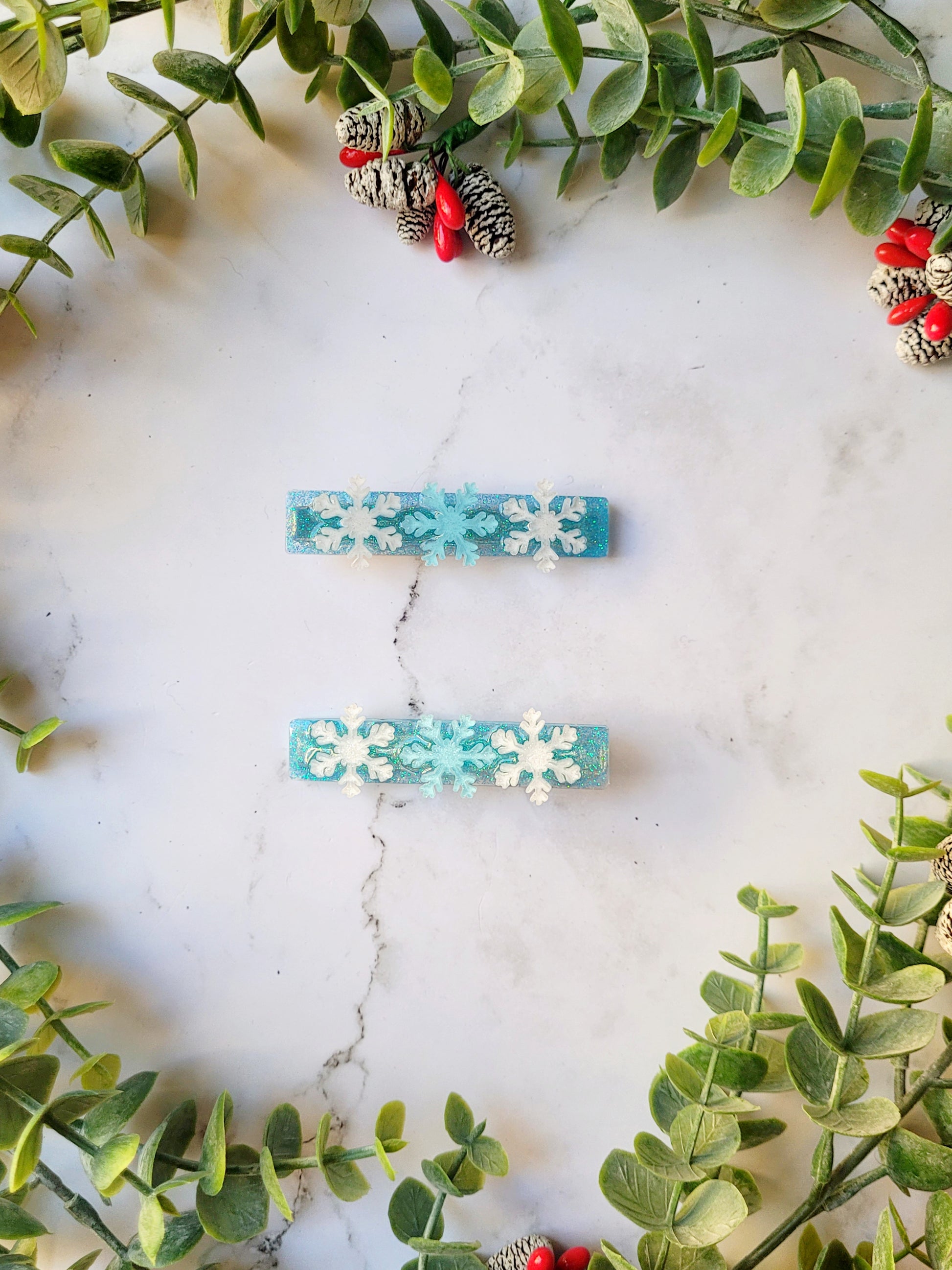 blue glitter hair clip with white and light blue snowflakes on a marble background with foliage. 