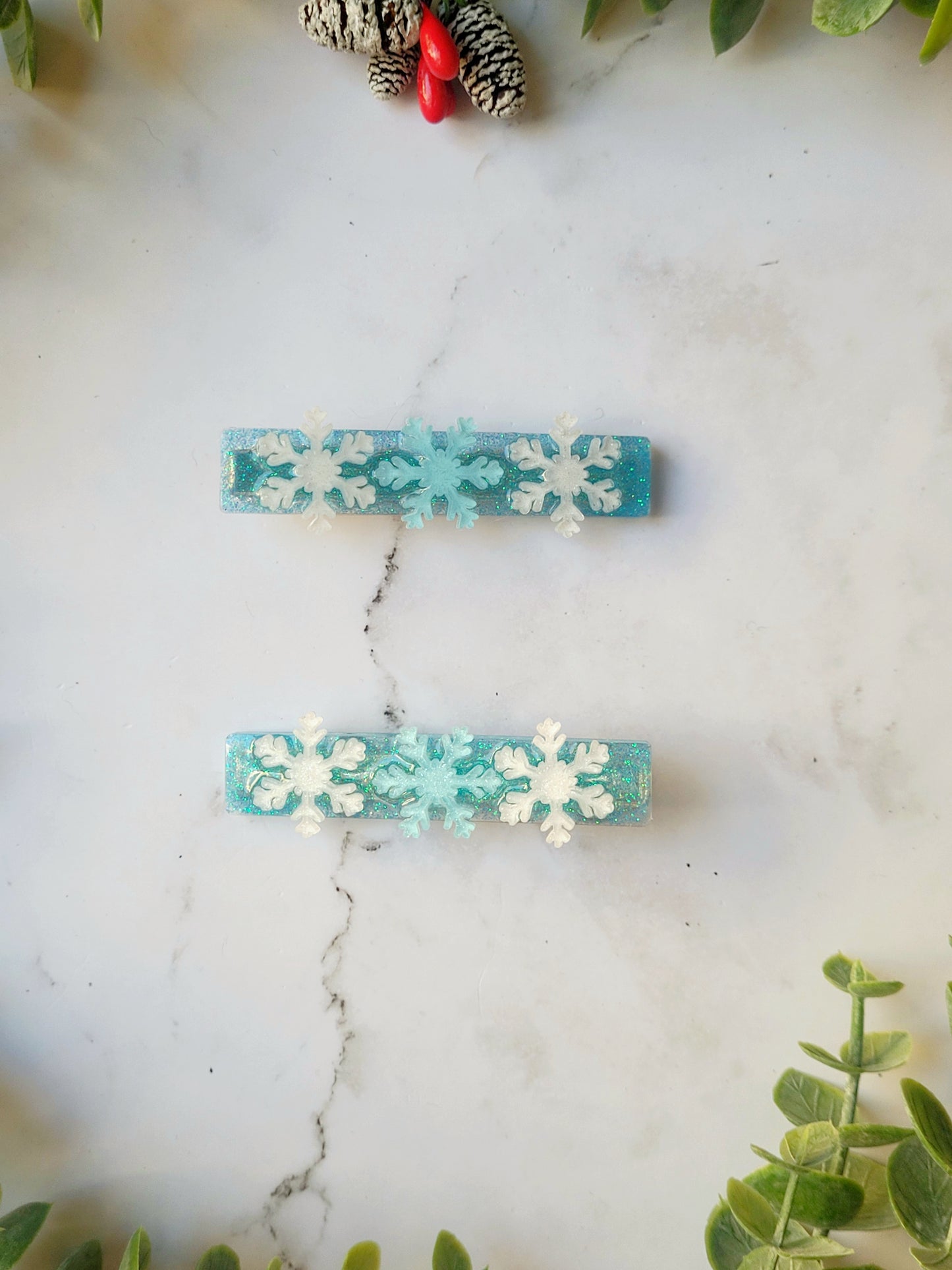 close up of blue glitter hair clip with white and light blue snowflakes on a marble background with foliage.