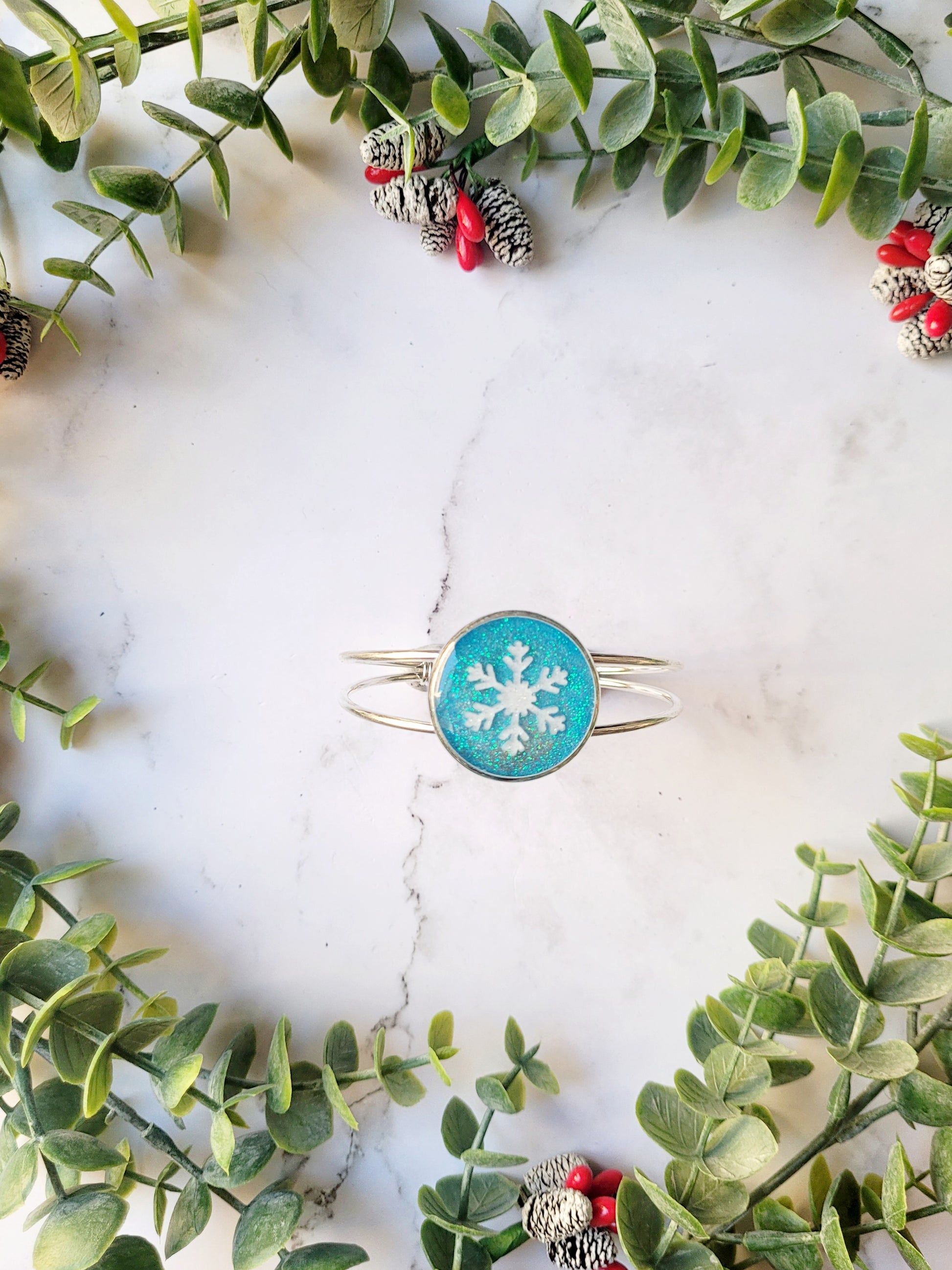 silver braceclet with a blue glitter charm and a white snowflake on  white marble background with foliage. 