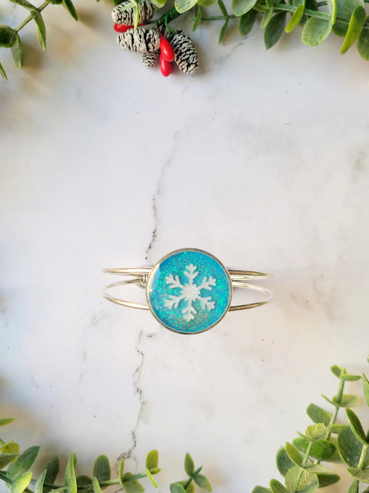 close up of silver braceclet with a blue glitter charm and a white snowflake on white marble background with foliage.