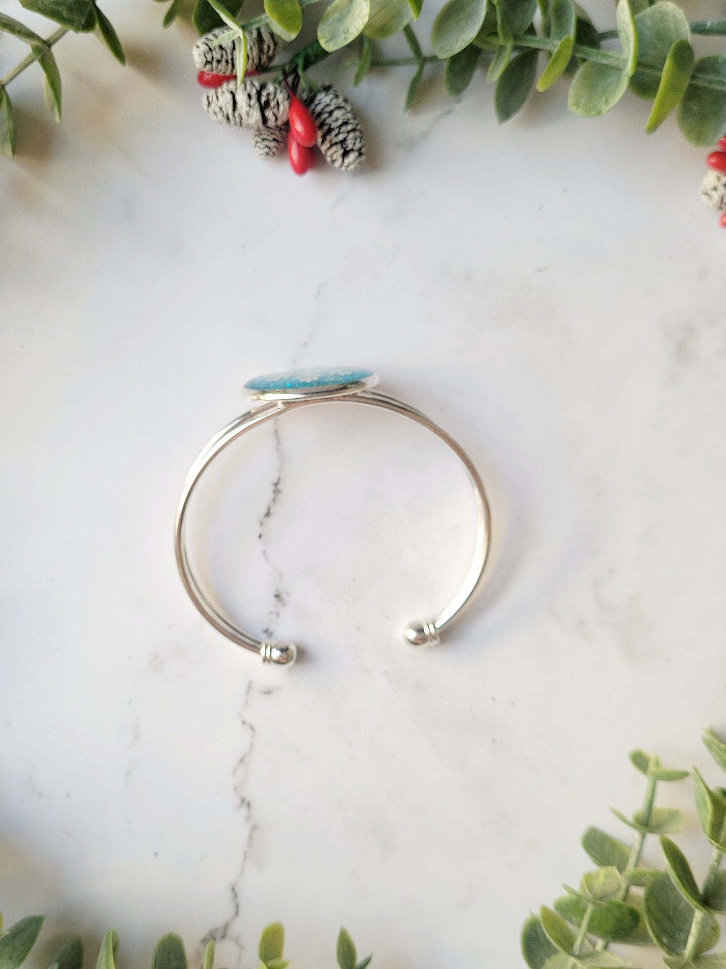 side view of silver braceclet with a blue glitter charm and a white snowflake on white marble background with foliage.