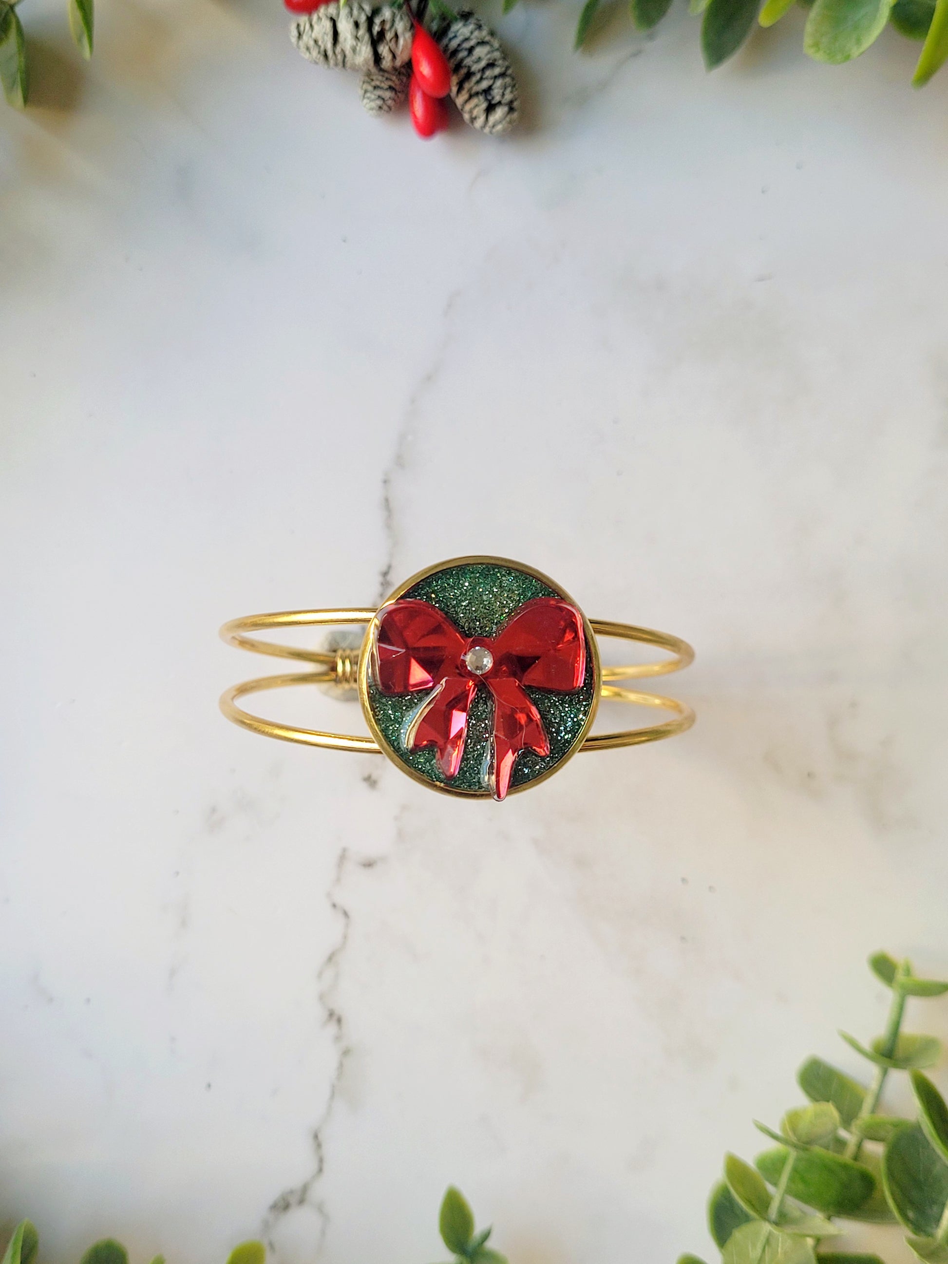 close up of green and red bow bracelet on a marble background surrounded by foliage.