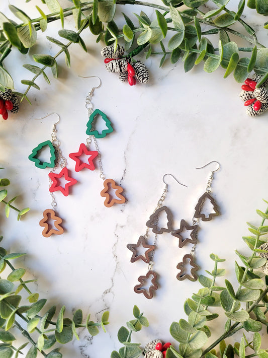Christmas cookie cutter dangle earrings in two color ways on a white marble background surrounded by foliage. 