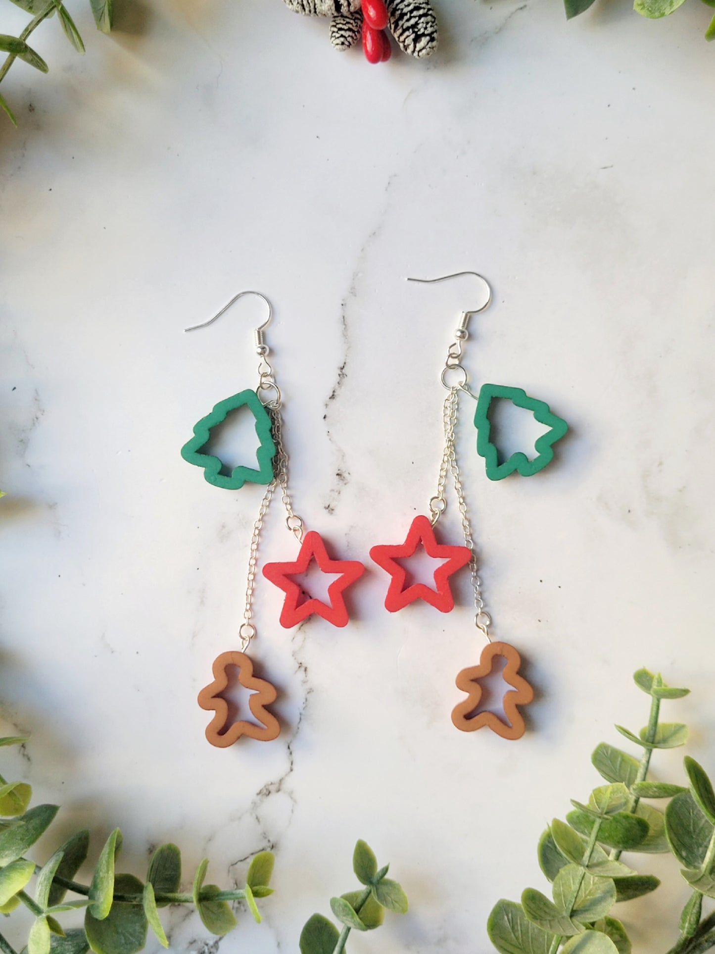 close up of Christmas cookie cutter dangle earrings in multi-color on a white marble background. .