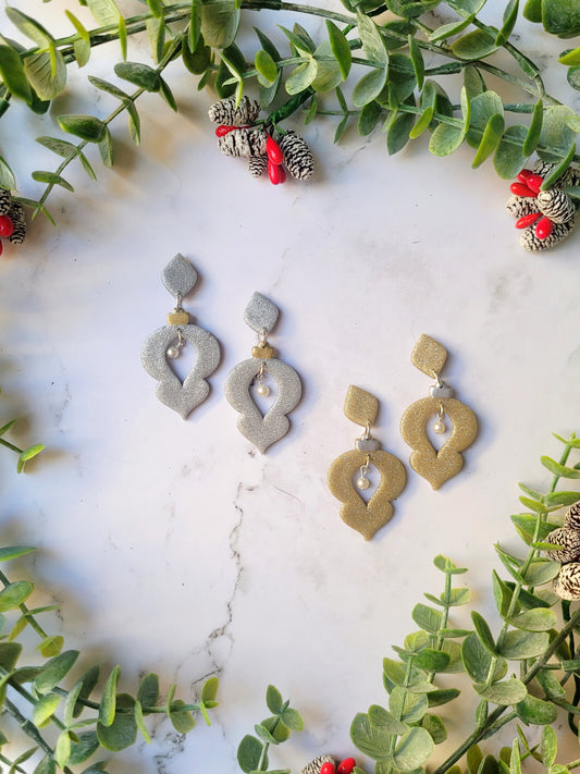 Gold and Silver glitter ornament shaped earrings with a pearl accent on a marble background with foliage. 