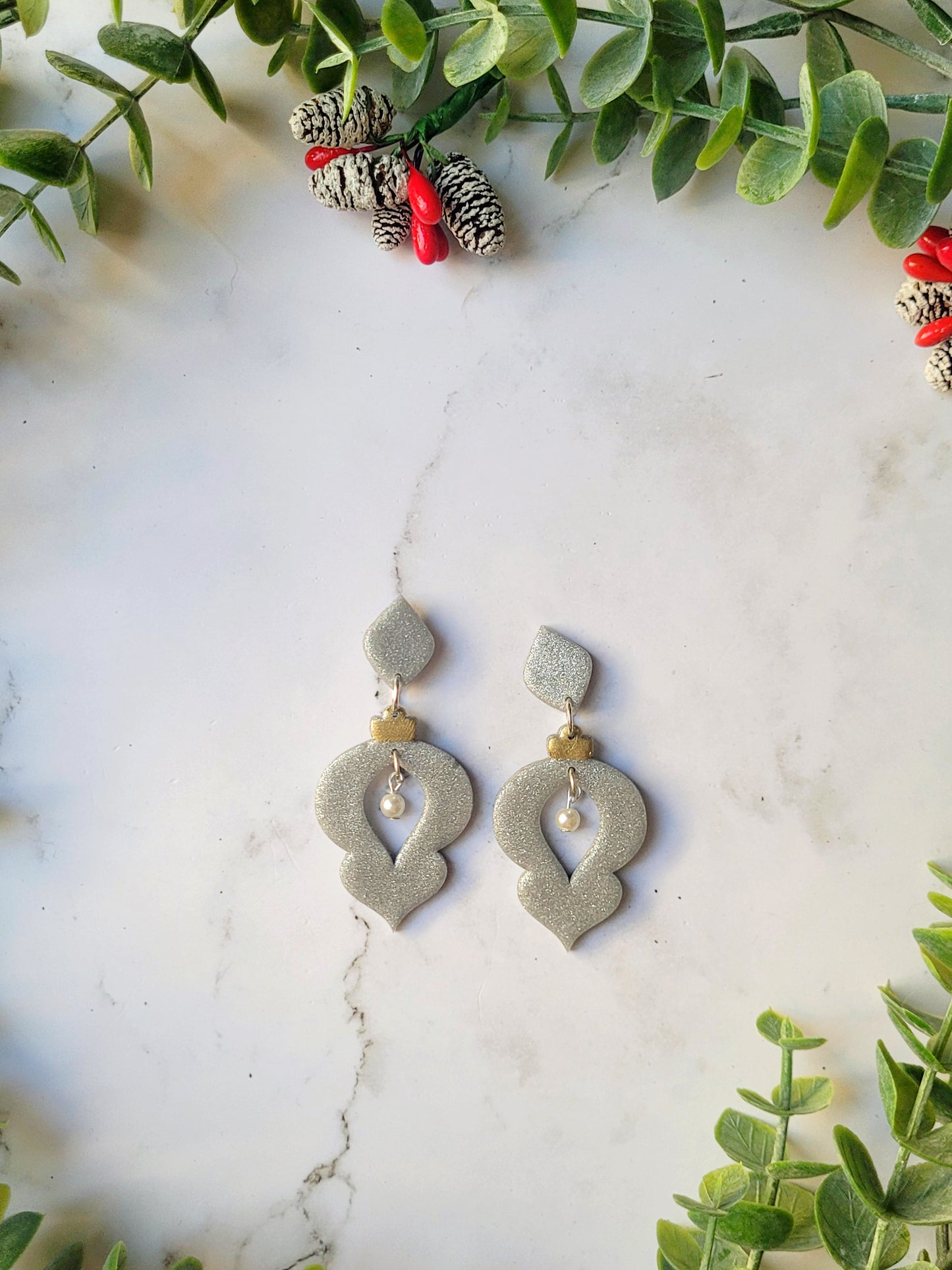 Silver glitter ornament shaped earrings with a pearl accent on a marble background with foliage. 