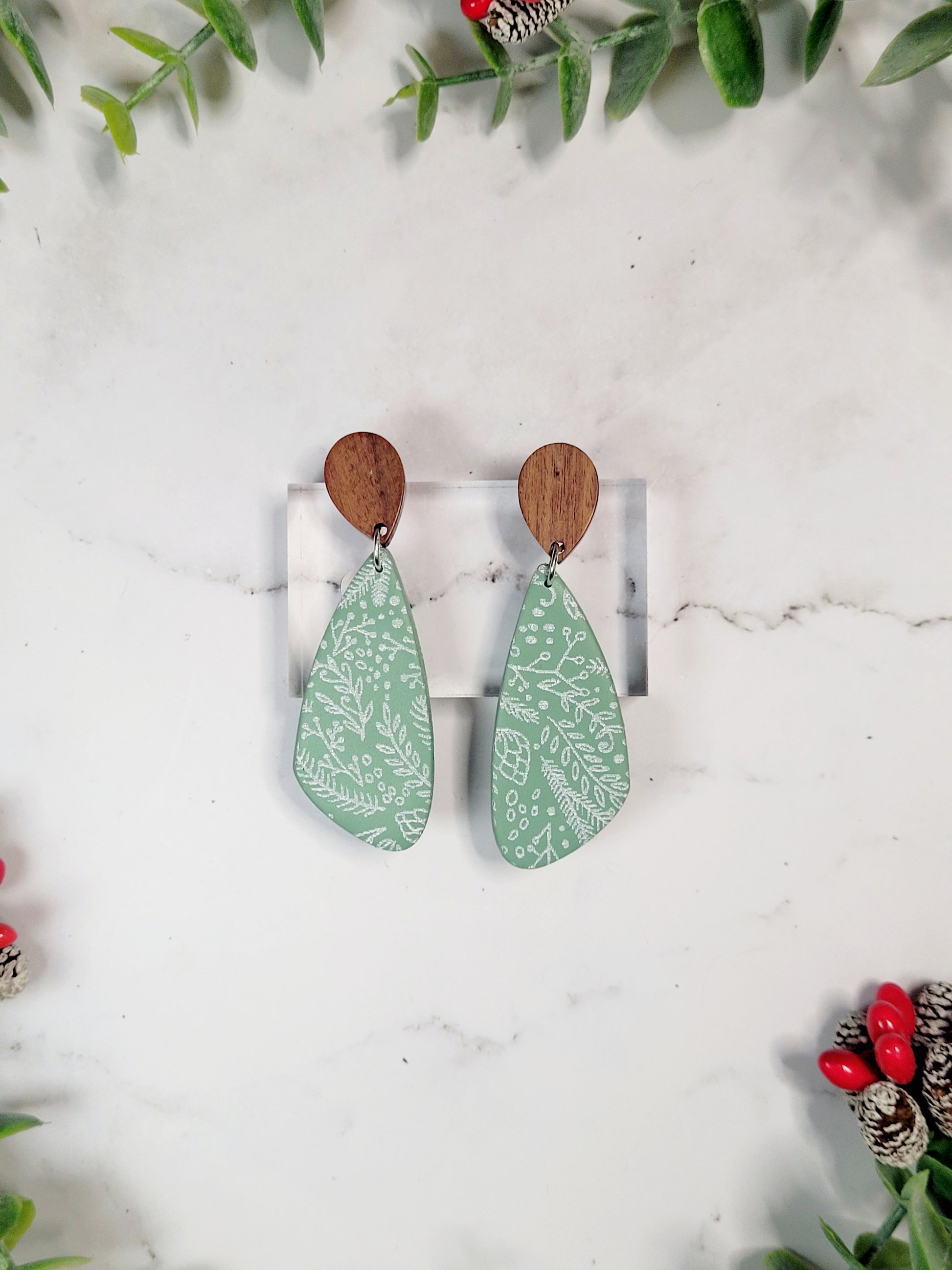 close up of mint green tear drop earrings with a winter botanical print and wood studs on a marble background with foliage.