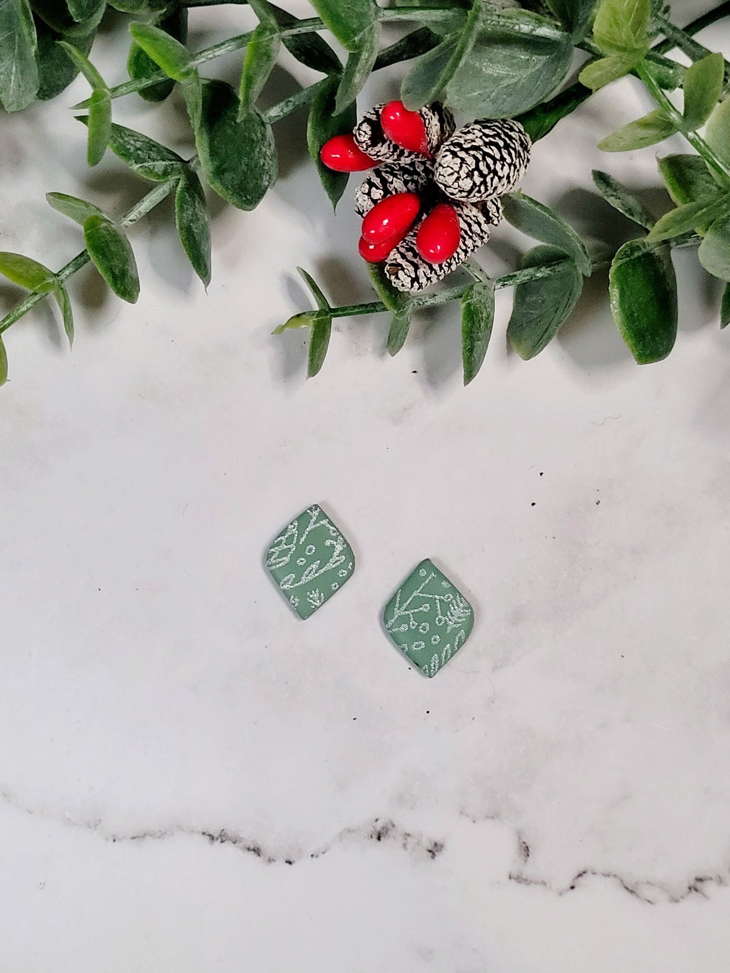 close up of mint green diamond shaped studs with a white botanical design on a marble background with foliage. 