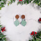 mint green winter botanical earrings on a marble background surrounded by foliage. 