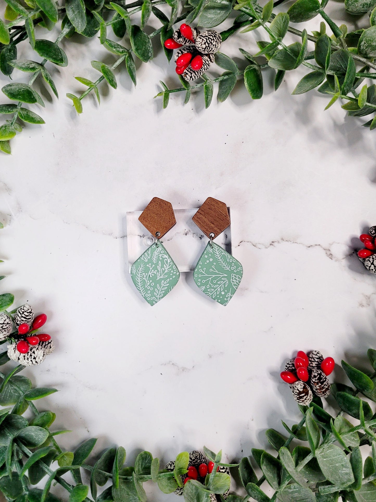 mint green winter botanical earrings on a marble background surrounded by foliage. 