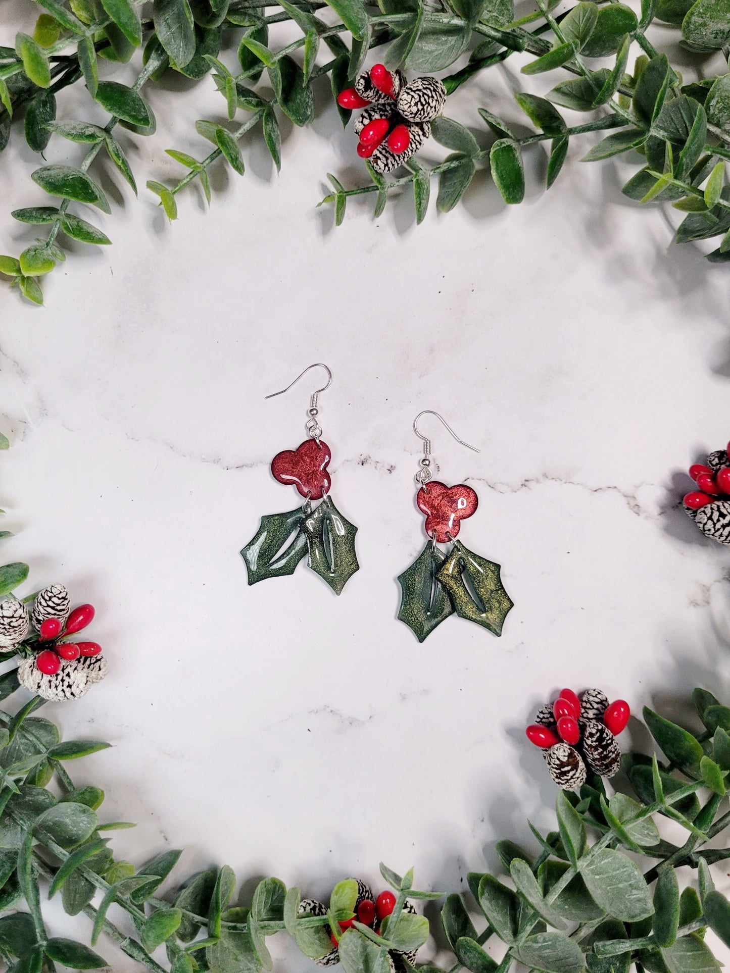 Glitter holly sprig earrings on a marble background surrounded by foliage, 