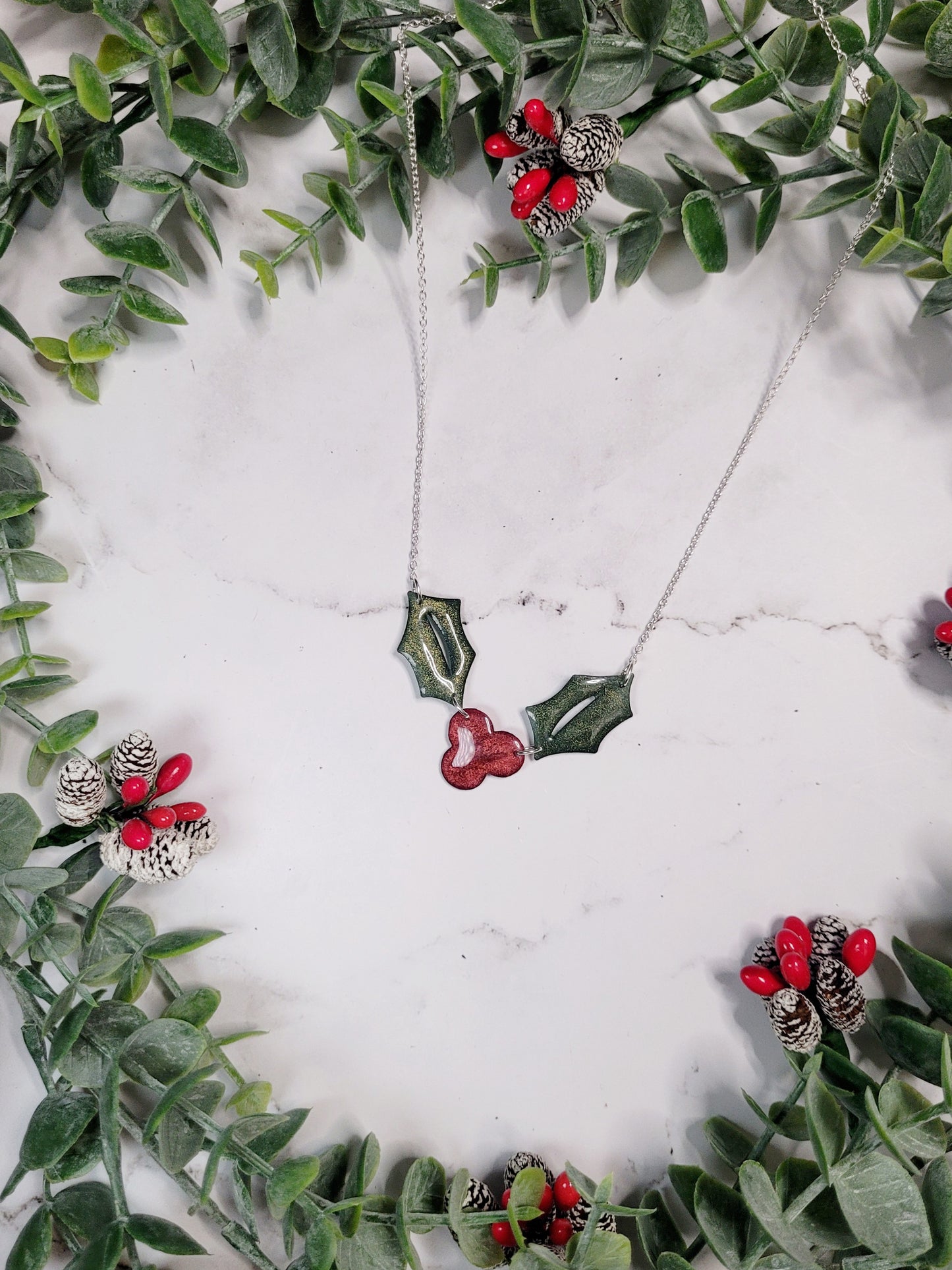 Glitter holly sprig necklace on a marble background surrounded by foliage,