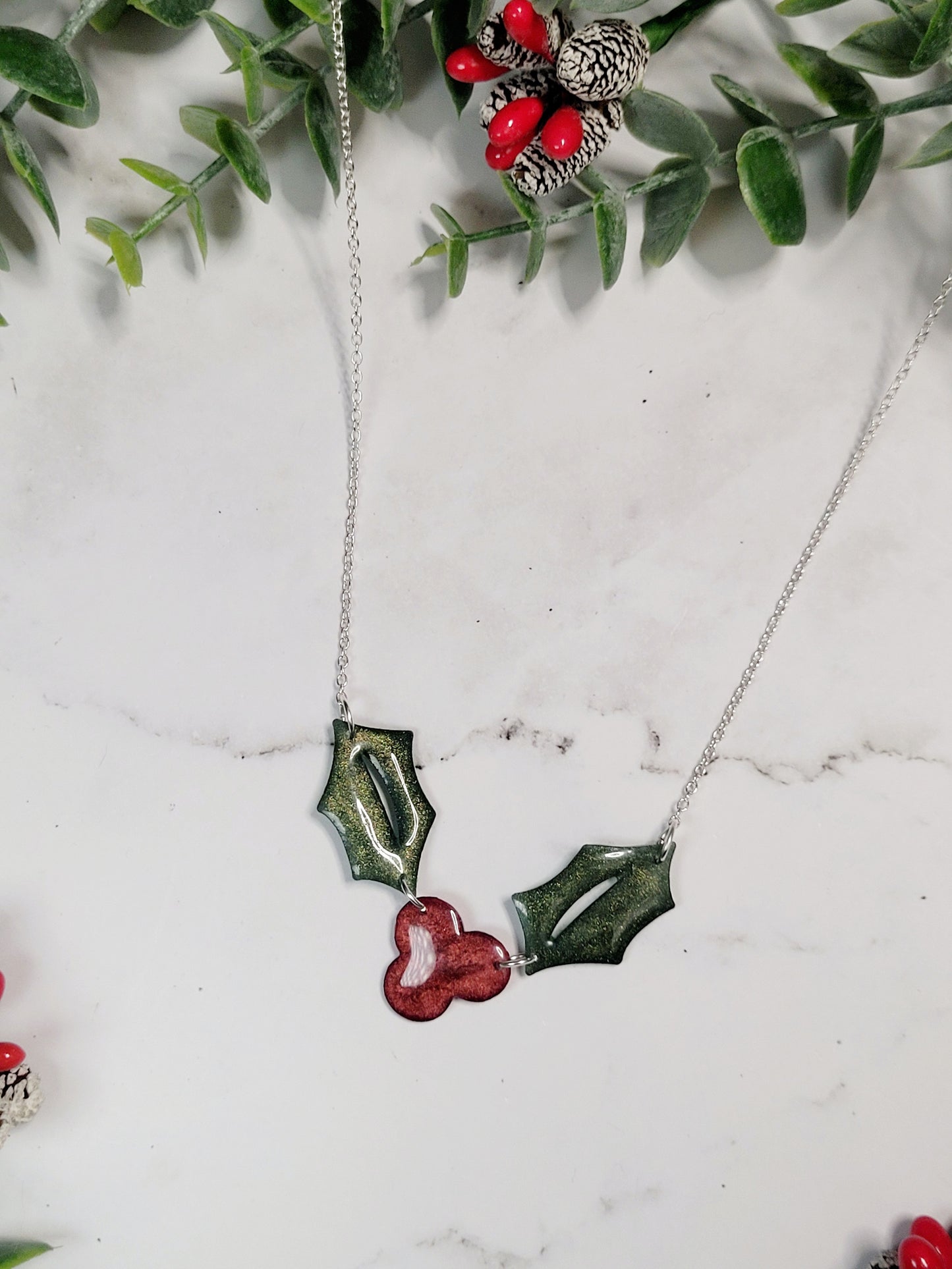 close up of Glitter holly sprig necklace on a marble background surrounded by foliage,