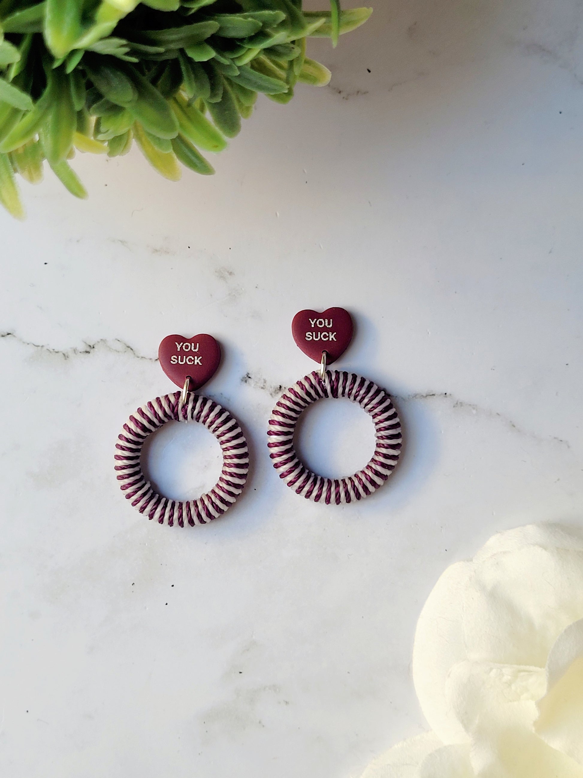 close up of burgundy and white striped hoop with a burgundy candy heart stud on a marble background with foliage. 