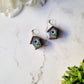the front of the 3-d blue birdhouse earrings on a white marble background surrounded by foliage.