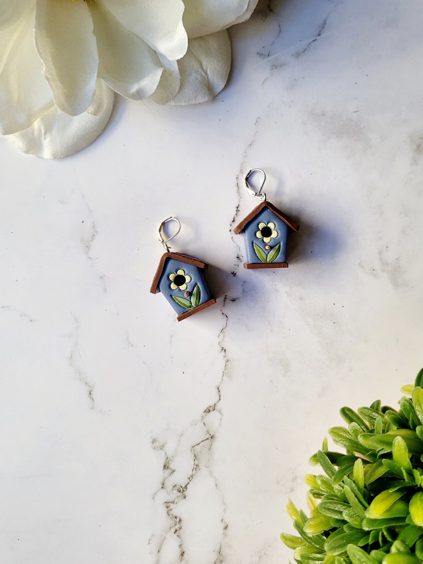 the front of the 3-d blue birdhouse earrings on a white marble background surrounded by foliage.