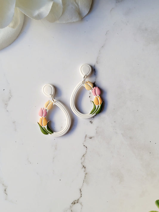 close up of white teardrop hoops with painted tulips on a marble background with foliage.