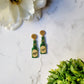 Glitter beer bottle shaped earrings on a white marble background surrounded by foliage. 