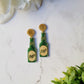 close up of Glitter beer bottle shaped earrings on a white marble background. 