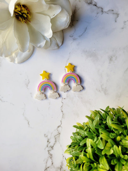 Rainbow dangle earrings with a star stud and cloud charms on a marble background with foliage. 