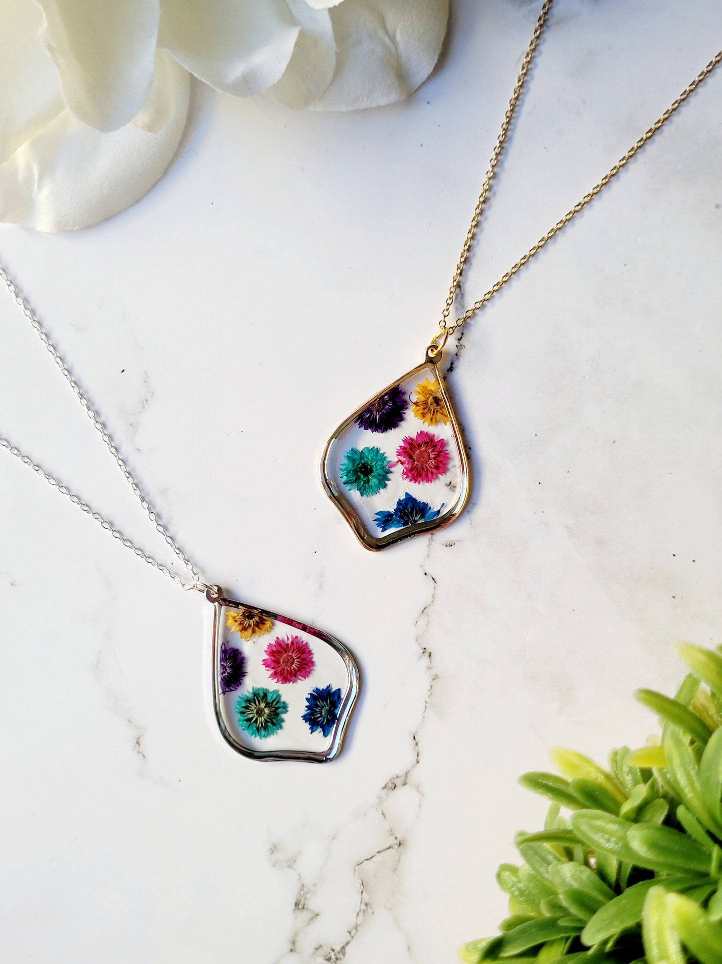close up of scalloped resin necklace with rainbow dried flowers on a marble background surrounded by foliage.
