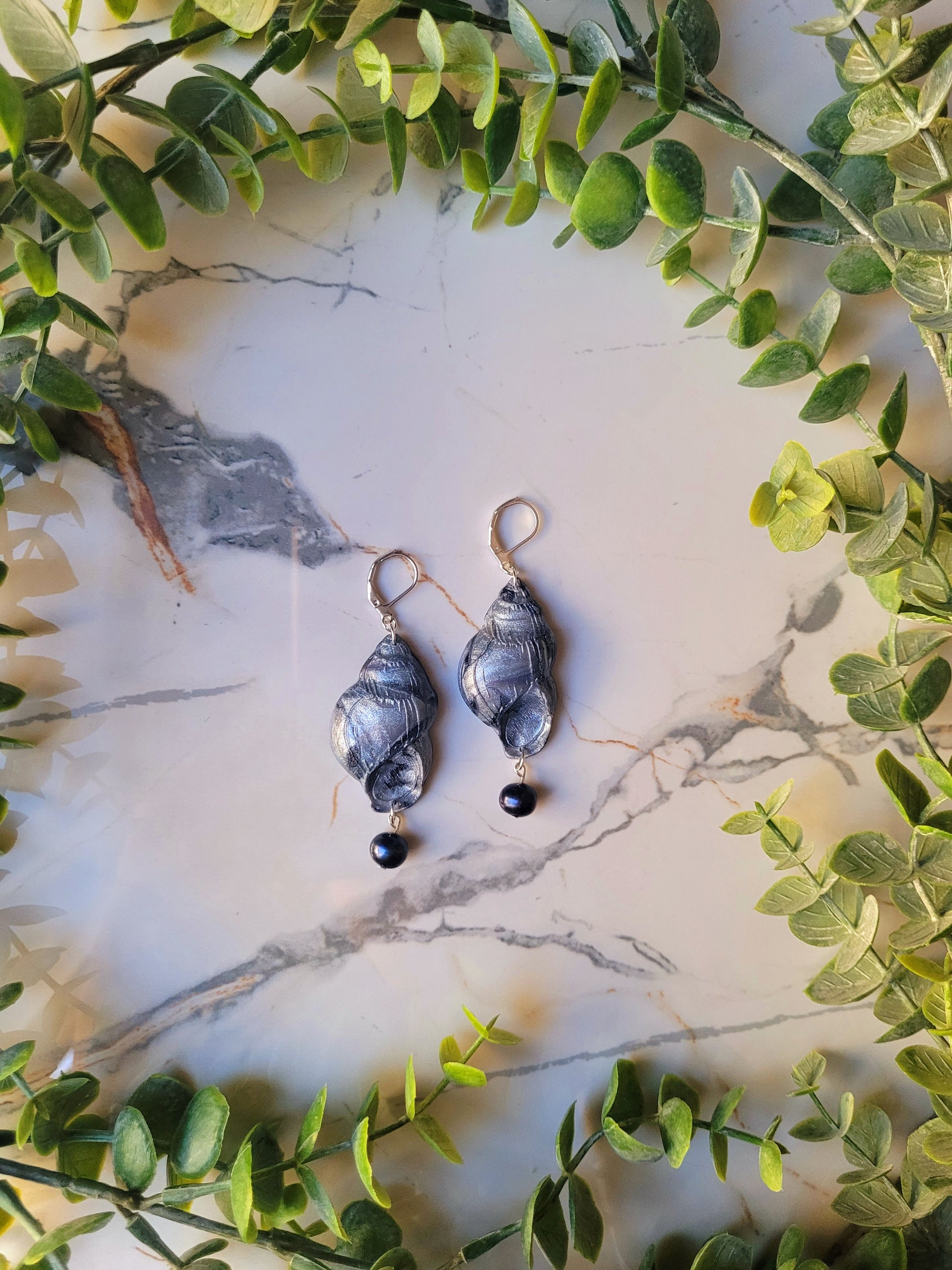 Dark silver conch shell with a black pearl on a marble background surrounded by foliage. 