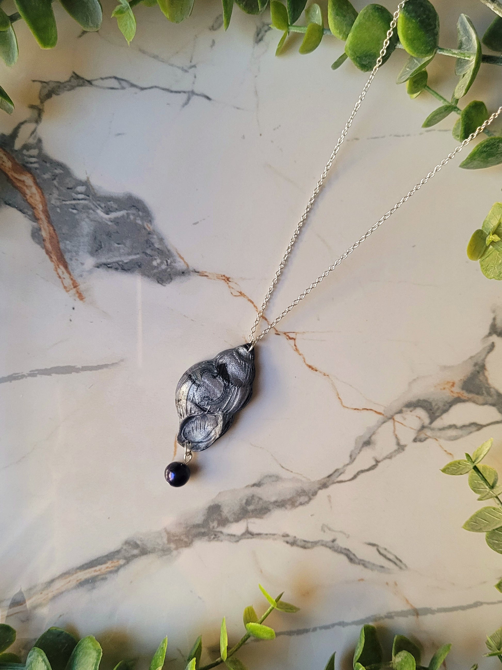 close up Dark silver conch shell with a black pearl necklace on a marble background surrounded by foliage.
