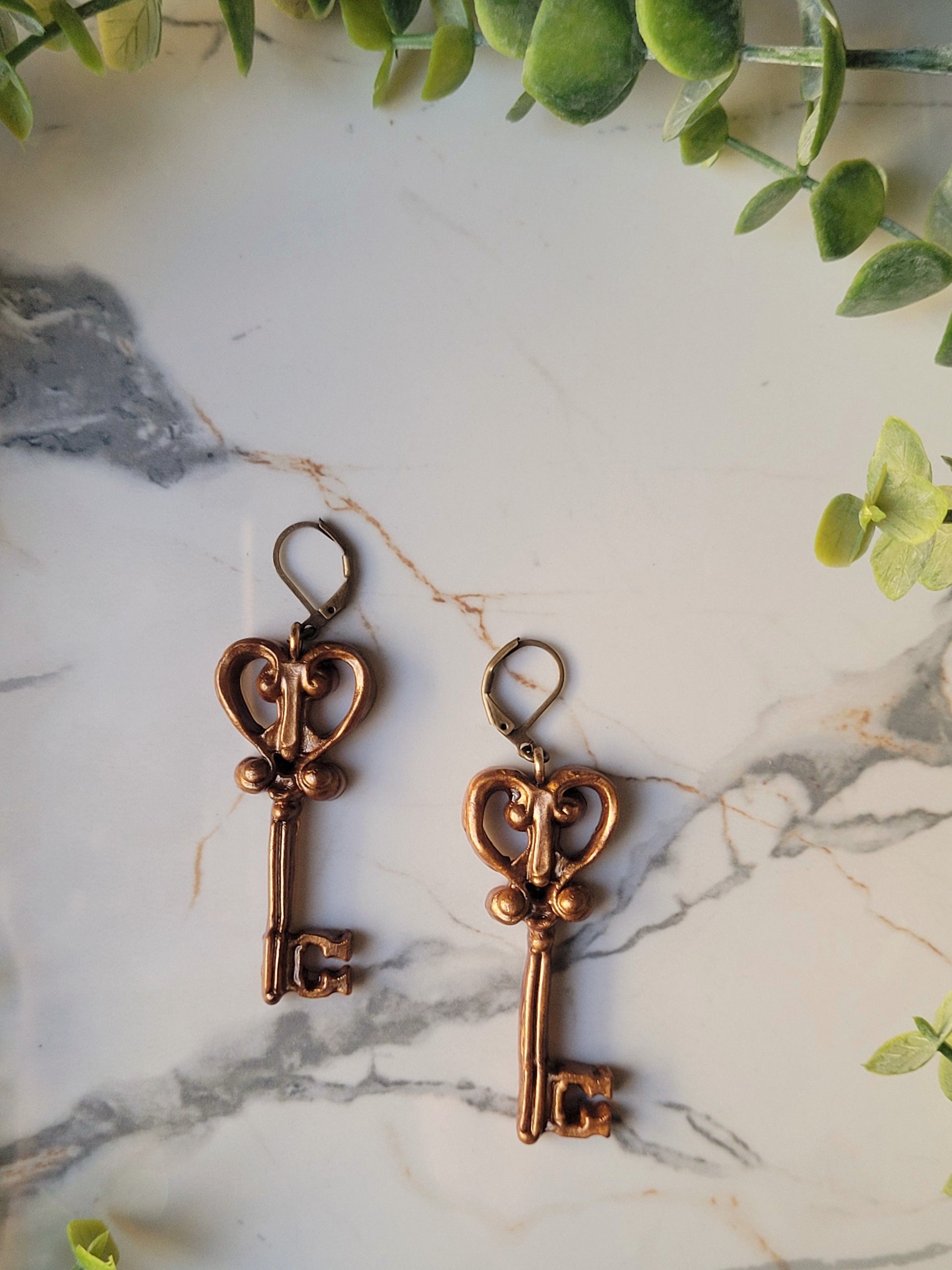 close up of heart skeleton key earrings with filigree in antique gold on a marble background with foliage. 