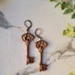 close up of spade shaped skeleton key earrings with filigree in antique gold on a marble background with foliage. 