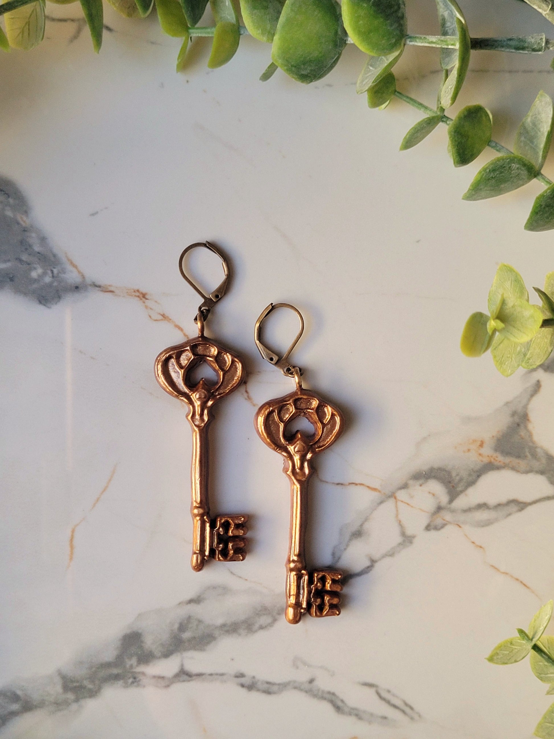 close up of spade shaped skeleton key earrings with filigree in antique gold on a marble background with foliage. 