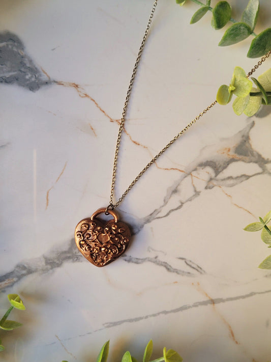 close up of heart shaped lock pendant with filigree in antique gold on a marble background with foliage. 