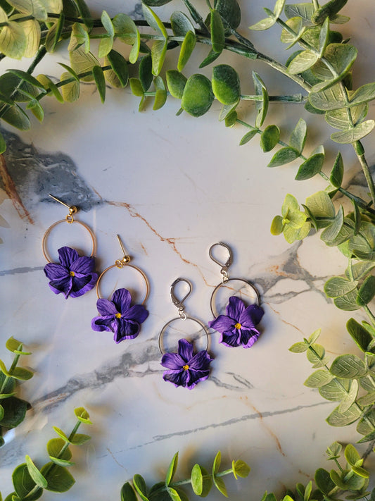 Purple pansy earrings with gold and silver findings on a marble background with foliage. 