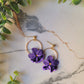 close up of the purple pansy earrings with gold findings on a marble background with foliage. 